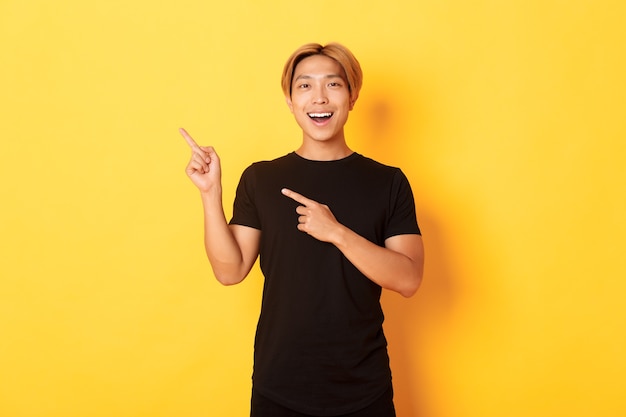 Ritratto di allegro uomo asiatico che punta le dita nell'angolo superiore sinistro, sorridente e mostrando soddisfatto, parete gialla.