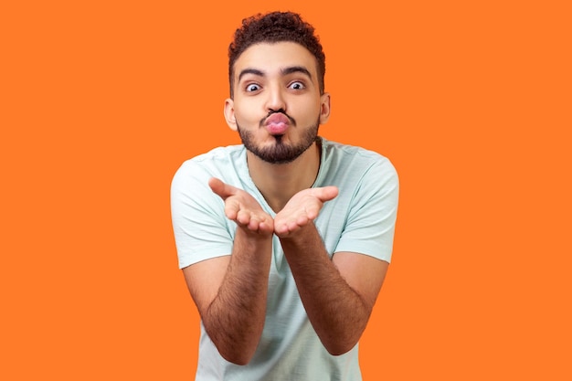 Ritratto di amoroso bell'uomo bruna con la barba in maglietta bianca casual che tiene i palmi in su mentre invia un bacio d'aria alla telecamera dimostrando sentimenti studio indoor girato isolato su sfondo arancione