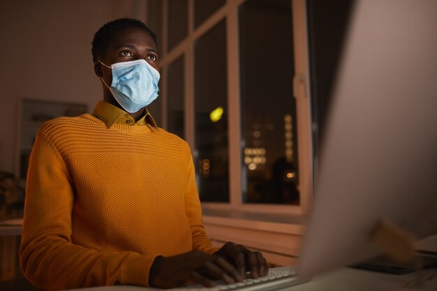 Ritratto di angolo basso di giovane uomo afro-americano che indossa la maschera in ufficio mentre si utilizza il computer illuminato dallo schermo nel buio, copia spazio