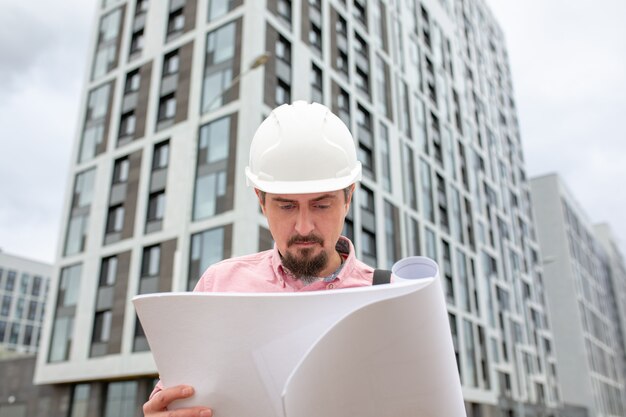 Ritratto di architetto al lavoro con casco in un cantiere edile, legge il piano, progetti di carta