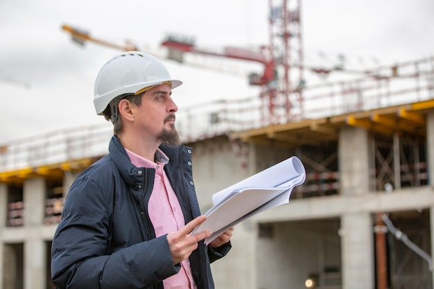 Ritratto di architetto al lavoro con casco in un cantiere edile, legge il piano, progetti di carta