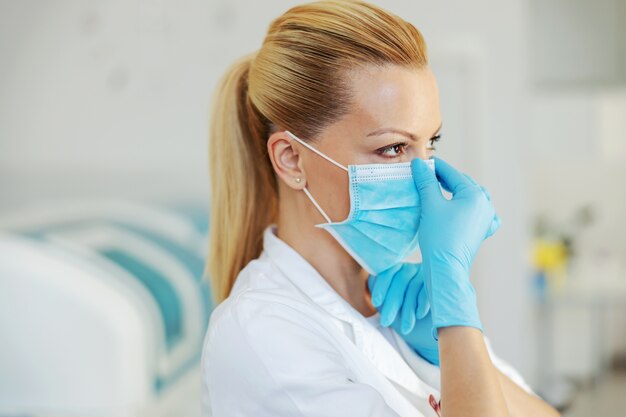 Ritratto di assistente di laboratorio bionda attraente con guanti di gomma in piedi in ospedale e mettendo su maschera facciale sterile.