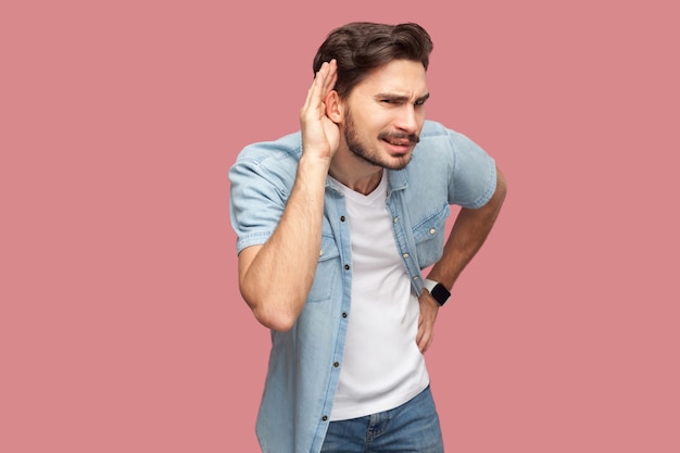 Ritratto di attento bel giovane barbuto in camicia blu stile casual in piedi con la mano sull'orecchio e cercando di sentire qualcosa con la faccia seria. girato in studio al coperto, isolato su sfondo rosa.