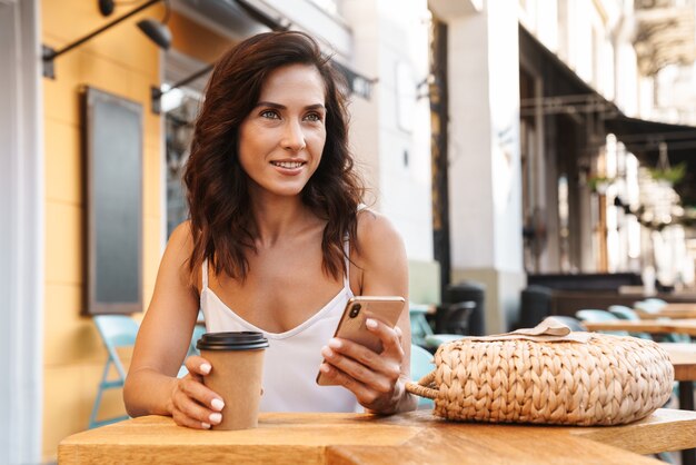 Ritratto di attraente donna bruna con borsa di paglia che beve caffè da un bicchiere di carta e usa il cellulare mentre è seduto in un accogliente bar all'aperto