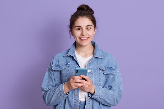 Ritratto di attraente donna caucasica indossa elegante giacca di jeans, in piedi al coperto contro il muro lilla con il moderno smartphone nelle mani,