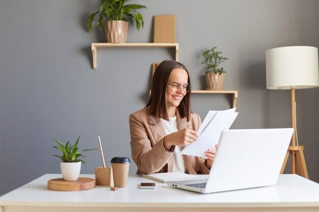 Ritratto di attraente donna soddisfatta sorridente con i capelli castani che indossa una giacca beige che lavora online su un computer portatile che tiene in mano documenti cartacei che si godono il suo lavoro