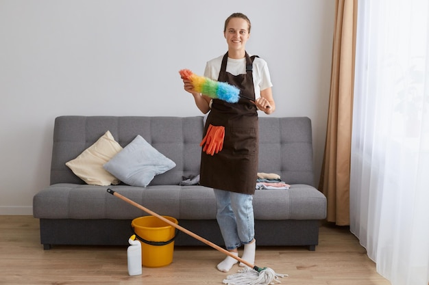 Ritratto di attraente donna sorridente che pulisce la sua casa indossando jeans e grembiule marrone casalinga che fa le sue faccende domestiche in piedi vicino alla tosse con ppduster colorato nelle mani