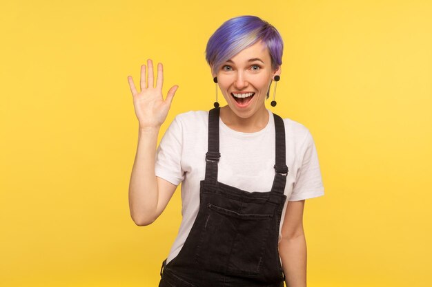Ritratto di attraente felice donna hipster amichevole con i capelli corti viola in tuta di jeans agitando la mano dicendo ciao salutando gli amici con ciao gesto isolato su sfondo giallo girato in studio