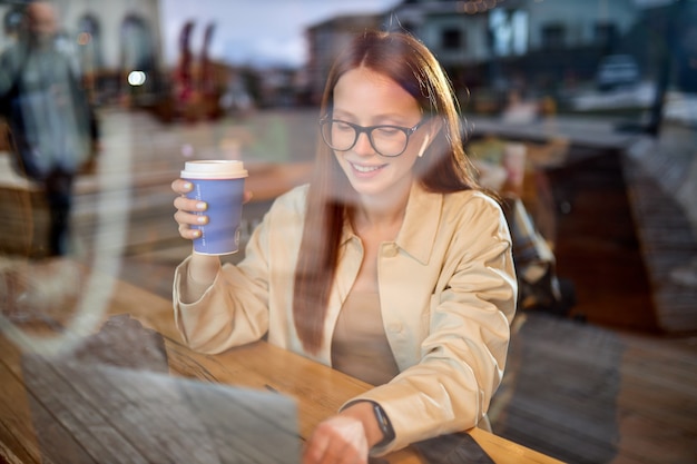 Ritratto di attraente giornalista fotografa in occhiali con i capelli rossi seduto al bar che lavora con computer netbook digitale, lavorando in remoto con file multimediali. foto attraverso la finestra