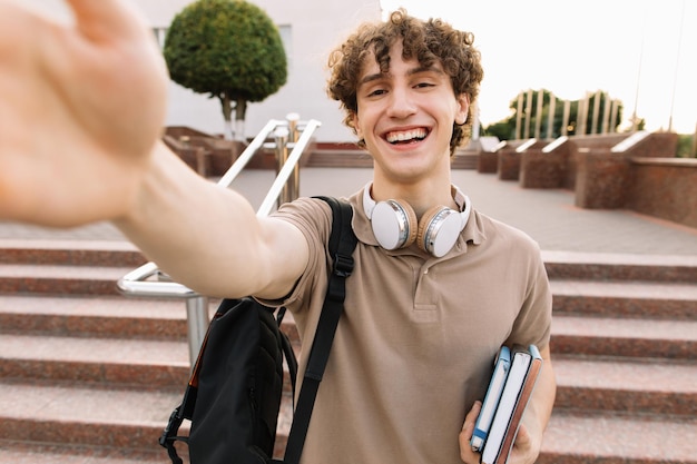 Ritratto di attraente studente maschio riccio in cuffie sorridente e prendendo il concetto di videochiamata selfie