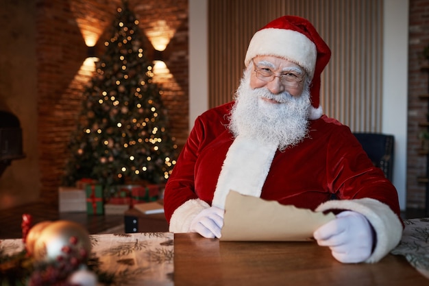 Ritratto di Babbo Natale anziano sorridente seduto al tavolo di legno in soggiorno e leggendo la lettera con...