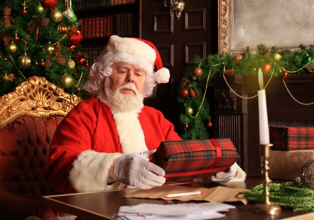 Ritratto di Babbo Natale felice seduto nella sua stanza a casa vicino all'albero di Natale con scatola regalo.