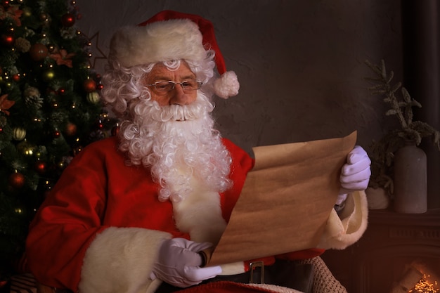 Ritratto di Babbo Natale felice seduto nella sua stanza a casa vicino all'albero di Natale e leggendo la lettera di Natale o la lista dei desideri.