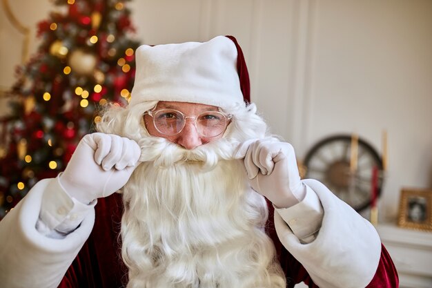 Ritratto di Babbo Natale in bicchieri toccando la sua barba vicino al camino e albero di Natale con doni.