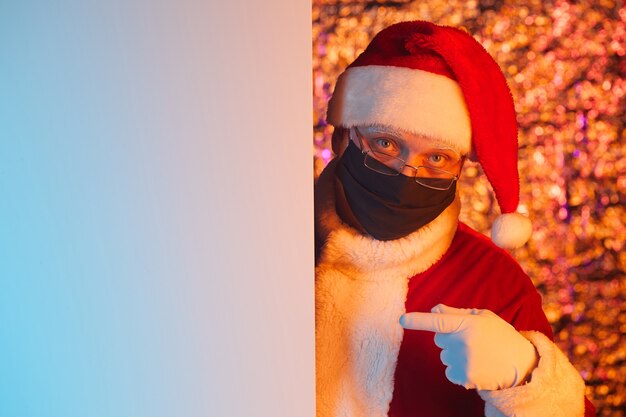 Ritratto di Babbo Natale in maschera protettiva che punta al poster vuoto che consiglia qualcosa