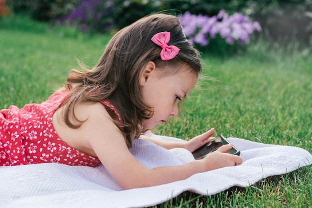 Ritratto di bambina 3-4 in rosso sdraiato su una coperta sull'erba verde e guardando nel telefono cellulare. Bambini che usano gadget