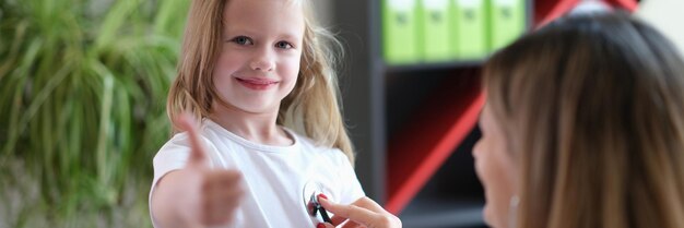 Ritratto di bambina all'appuntamento dal medico nell'ufficio della clinica che mostra il pollice sulla donna del segno
