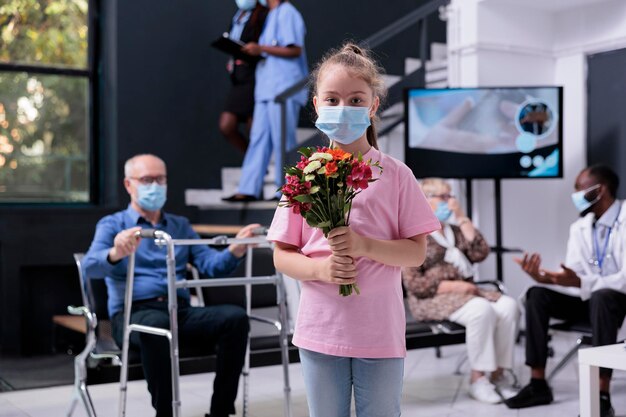 Ritratto di bambina che indossa una maschera facciale di protezione medica per prevenire l'infezione da covid19, in piedi nell'area di attesa dell'ospedale. Bambino che tiene il mazzo di fiori per la nonna. Servizio di medicina