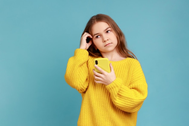 Ritratto di bambina che tiene in mano lo smartphone, distogliendo lo sguardo con un'espressione facciale premurosa, indossando un maglione giallo in stile casual. Studio indoor girato isolato su sfondo blu.