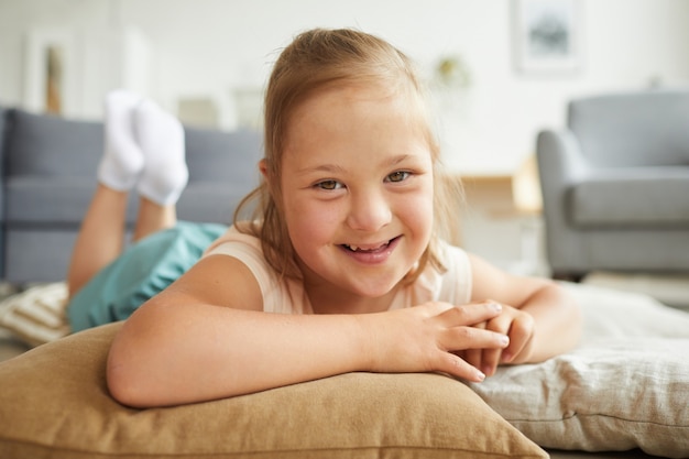 Ritratto di bambina con sindrome di down sorridere alla telecamera mentre giaceva sui cuscini sul pavimento