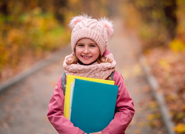 Ritratto di bambina con zaino tenendo le cartelle mentre si sta in piedi sulla strada del parco in autunno