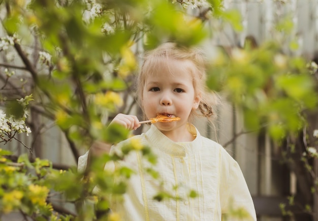 Ritratto di bambina divertente in abito giallo che succhia caramelle sullo sfondo giardino fiorito