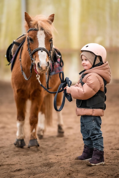 Ritratto di bambina in giacca protettiva e casco con il suo pony marrone prima della lezione di equitazione