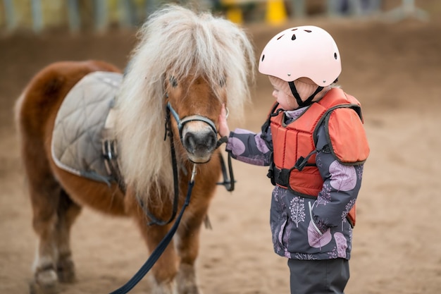 Ritratto di bambina in giacca protettiva e casco con il suo pony marrone prima della lezione di equitazione