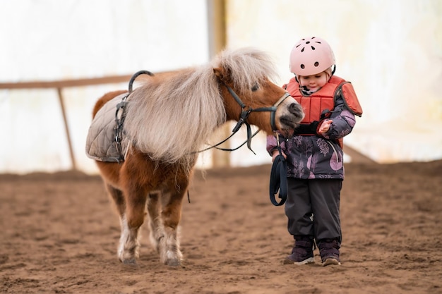 Ritratto di bambina in giacca protettiva e casco con il suo pony marrone prima della lezione di equitazione