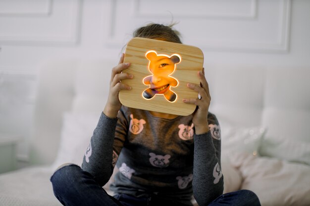 Ritratto di bambina seduta sul letto nella posizione del loto e guardando attraverso l'elegante lampada da notte in legno con foto di orso.