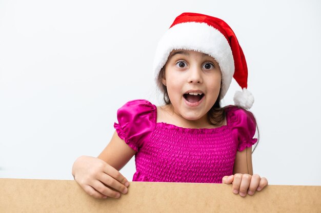 Ritratto di bambina sorridente con cappello di Natale. Bambino che fa capolino dietro la scheda vacanze vuota. Bambino carino felice in posa dietro il pannello di carta kraft, isolato su sfondo bianco.