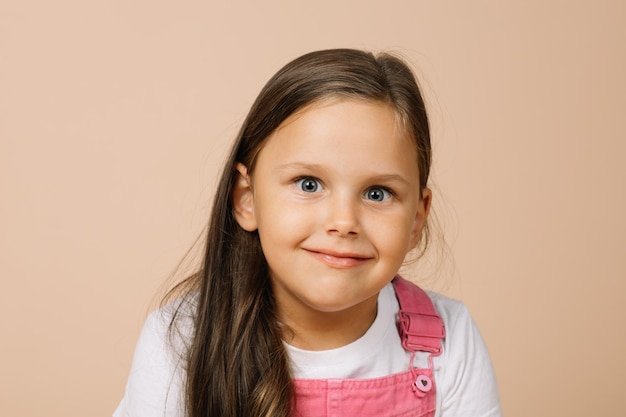 Ritratto di bambino con occhi luminosi leggermente sporgenti e sorriso beato guardando la fotocamera che indossa una tuta rosa brillante e una maglietta bianca su sfondo beige