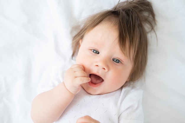 Ritratto di bambino divertente con il dito in una bocca sorridente e sdraiato su una biancheria da letto bianca a casa.