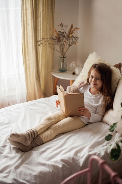 Ritratto di bambino felice La piccola bella ragazza riccia abbraccia il suo coniglio di peluche e legge il libro nel suo letto Casa accogliente