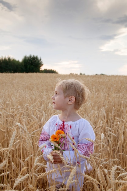 Ritratto di bambino in camicia con ricamo rosso e bouquet di fiori in mano nel campo di grano al tramonto