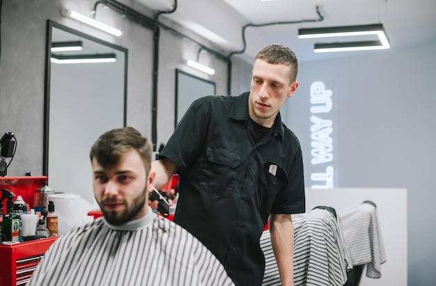 Ritratto di barbiere professionista che lavora sul taglio di capelli di un uomo adulto con la barba in un negozio di barbiere leggero