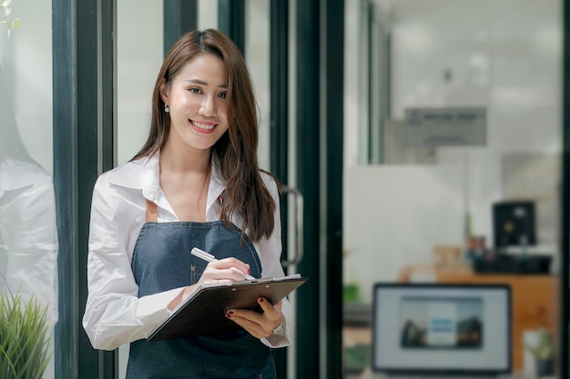 Ritratto di barista femminile fiducioso in piedi davanti al bancone. Proprietario del caffè della donna in grembiule che guarda l'obbiettivo e sorridente.