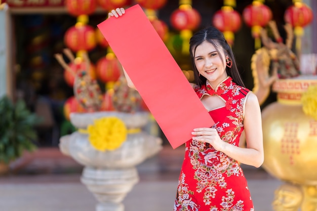 Ritratto di bei sorrisi giovane donna asiatica che indossa rosso cheongsam cinese tradizionale, che tiene mostra carta bianca primavera rossa per il Festival di Capodanno cinese al santuario cinese