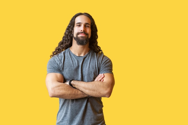 Ritratto di bel giovane barbuto soddisfatto con lunghi capelli ricci in maglietta grigia in piedi con le braccia incrociate e guardando la telecamera con un sorriso. studio al coperto colpo isolato su sfondo giallo.