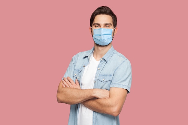 Ritratto di bel giovane con maschera medica chirurgica in camicia blu stile casual in piedi, braccia alzate e guardando la telecamera con un sorriso. girato in studio al coperto, isolato su sfondo rosa.