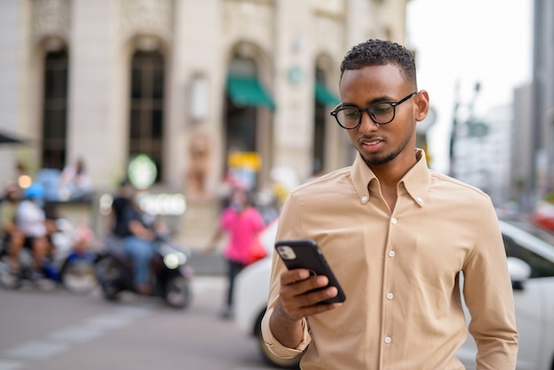 Ritratto di bel giovane uomo d'affari africano nero che indossa abiti casual all'aperto in città e utilizzando il telefono cellulare