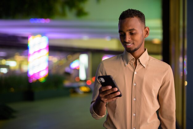 Ritratto di bel giovane uomo d'affari africano nero che indossa abiti casual all'aperto in città e utilizzando il telefono cellulare