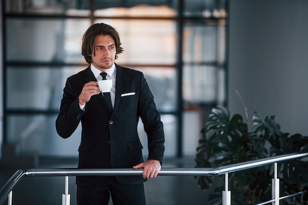Ritratto di bel giovane uomo d'affari in abito nero e cravatta si trova al chiuso con una tazza di bevanda.