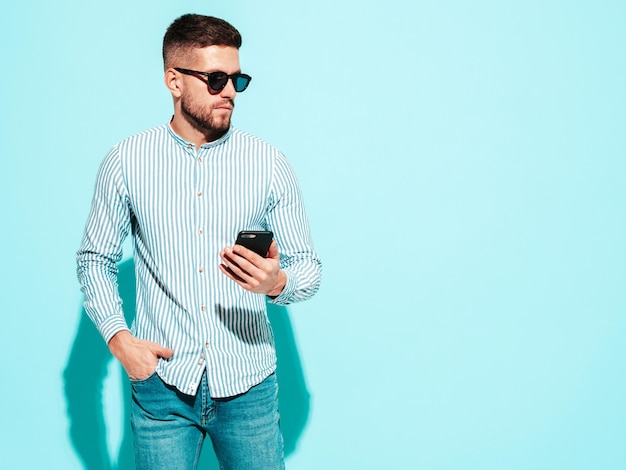 Ritratto di bel modello fiducioso Uomo alla moda sexy vestito in camicia e jeans Moda uomo hipster in posa vicino al muro blu in studio in occhiali da sole Holding smartphone