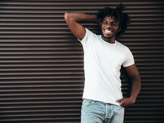 Ritratto di bel modello hipsterUnshaven africano uomo vestito con maglietta bianca estiva e jeans Moda maschile con acconciatura di dreadlocks in posa vicino al muro della tapparella in strada