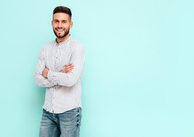 Ritratto di bel modello sorridente Uomo alla moda sexy vestito in camicia e jeans Moda uomo hipster in posa vicino al muro blu in studio Braccia incrociate