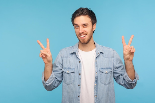 Ritratto di bell'uomo positivo che indossa una camicia di jeans che mostra il segno di v gesto di vittoria con le dita che guardano la fotocamera con un sorriso a trentadue denti Colpo di studio al coperto isolato su sfondo blu