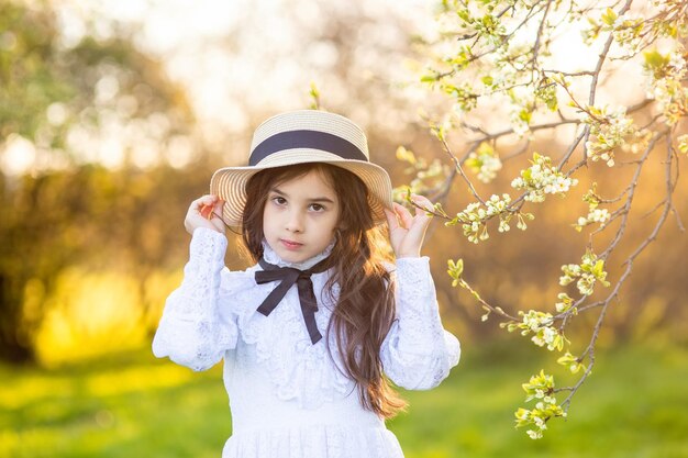 Ritratto di bella bambina con cappello e abito bianco in piedi sotto gli alberi in fiore in primavera