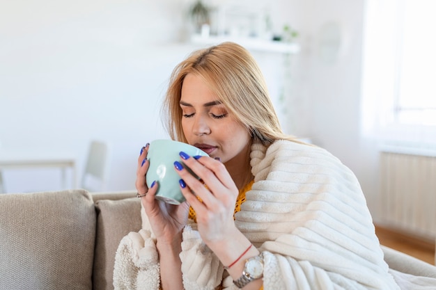Ritratto di bella bella giovane donna che beve caffè caldo a casa - al chiuso coperto con una coperta che tiene una tazza di tè, seduta sul divano, divano
