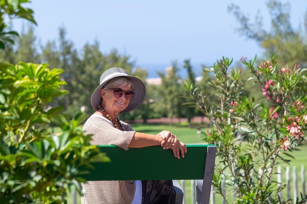 Ritratto di bella donna anziana che indossa berretto e occhiali seduto all'aperto su una panchina del parco pubblico guardando la fotocamera Donna anziana sorridente che si diverte a rilassarsi in una giornata di sole